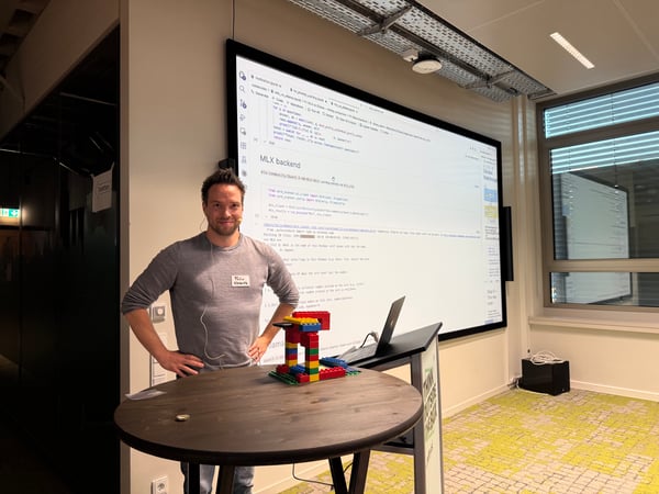 A man stands next to a table with a Lego structure, presenting in front of a large screen displaying Python code and text about a Pokémon-related AI project.