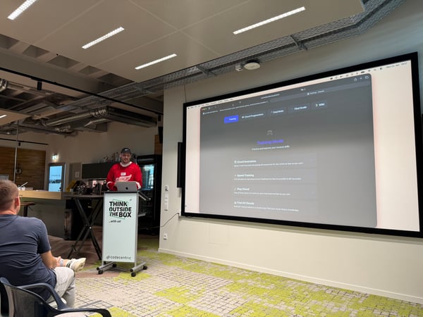A man in a red jersey is giving a presentation in a modern office setting, using a large screen displaying a music training application while an audience member watches.