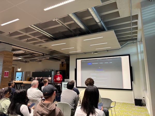 A speaker in a red jersey gives a presentation to an audience in a modern office space, with a large screen displaying a music theory training website.