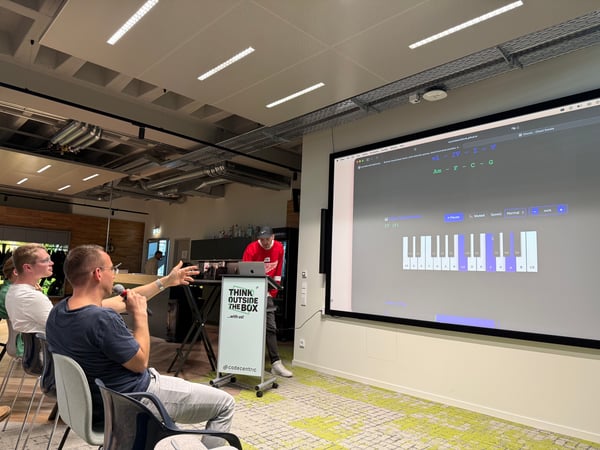 A man holds a microphone and points towards a large screen displaying a music application during a presentation in a modern office setting.