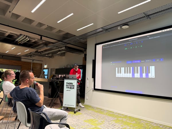 A man in a red jersey is giving a presentation about a music application on a large screen to a seated audience in a modern office space.