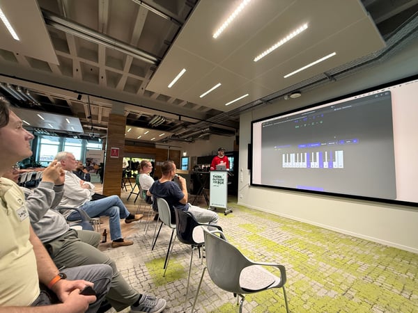 A group of people in a modern indoor space are watching a presentation about music theory or coding on a large screen.