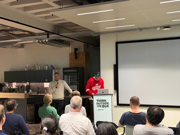 A man stands at the front of a room speaking into a microphone during a presentation, while another man works at a laptop behind a podium labeled 'THINK OUTSIDE THE BOX'.
