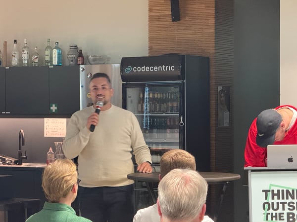 A man in a beige sweater is speaking into a microphone during an event in an office setting, with a 'codecentric' branded refrigerator in the background.