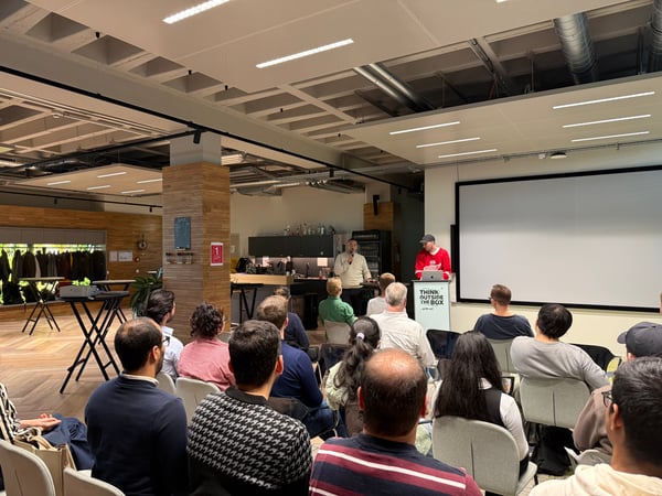 A group of people are gathered in a modern office space for a presentation, with two speakers standing at the front near a large projector screen.