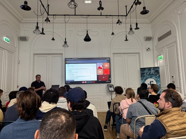 A photograph captures a man giving a technical presentation to a large audience seated in an ornate, high-ceilinged white room, with the presentation content displayed on a large central screen.