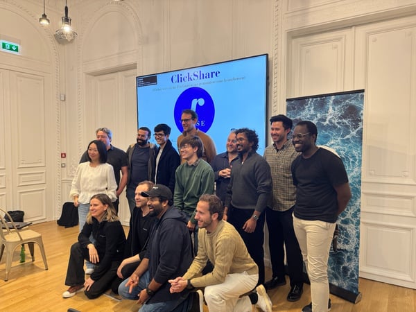 A group of thirteen people are posing for a team photo indoors in a large, ornate room with high white ceilings and a wooden floor. Behind the diverse group is a large screen displaying the 'ClickShare' logo and text, next to a tall roll-up banner featuring a turbulent blue water pattern.