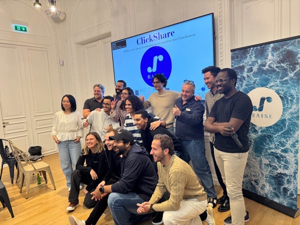 A group photo capturing approximately fourteen people, likely colleagues or a team, posing cheerfully indoors in an ornate event space. In the background, a large screen displays the 'ClickShare' logo and promotional text, positioned next to a vertical banner featuring the 'RAISE' logo against a blue, water-textured background.
