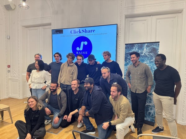 A group of fourteen people, likely colleagues or event attendees, pose for a group photo indoors in front of a large screen displaying the 'ClickShare' and 'RAISE' logos and French text.