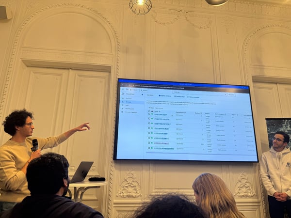 A technical presentation is taking place indoors, showing a speaker pointing at a large screen displaying the Google Cloud Run console interface with a list of services. Several audience members are visible in the foreground, and another presenter stands to the right against an ornate white wall.
