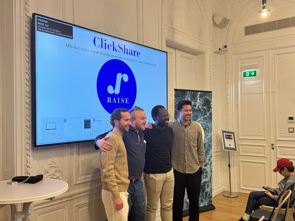 A photograph captures four men standing shoulder-to-shoulder and smiling in front of a large digital screen displaying presentation content related to 'ClickShare' and 'RAISE' in a formal, ornate indoor setting. The screen shows setup instructions primarily in French.