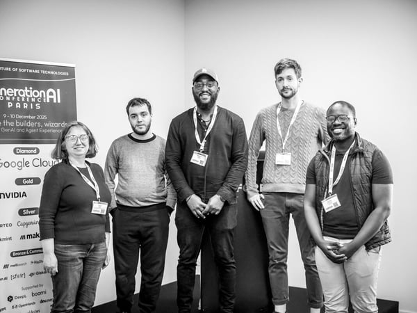 A black and white photograph captures five people—one woman and four men—standing together indoors in front of a conference banner listing sponsors and event details.