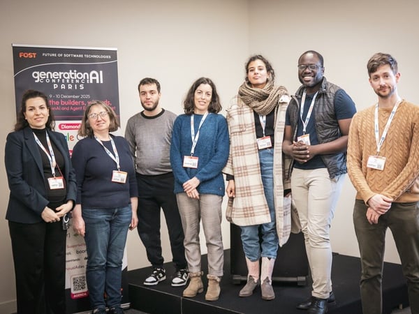 A group photograph featuring seven individuals, four women and three men, standing side-by-side on a small stage indoors at a conference or event, with a black and white promotional banner visible on the left.