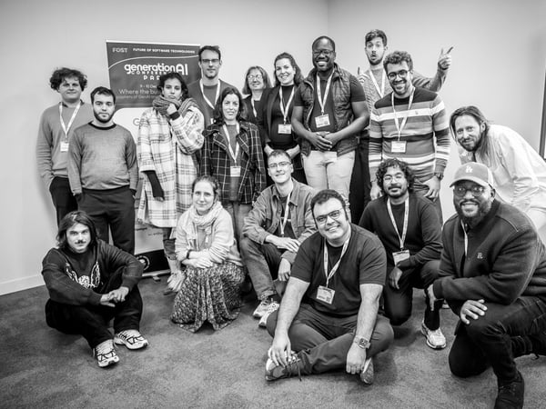 A black and white group photograph captures approximately sixteen people, likely attendees of a technology conference, posing indoors in front of a promotional banner.