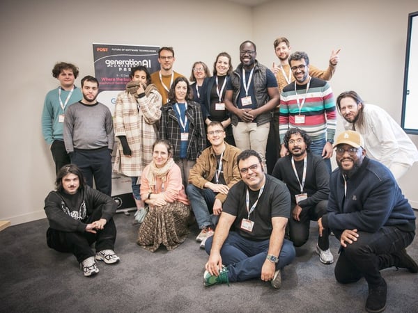A diverse group of approximately fifteen people, likely attendees or participants of a tech conference, pose for a group photo indoors in front of an event banner and a display screen showing 'WINNERS'.