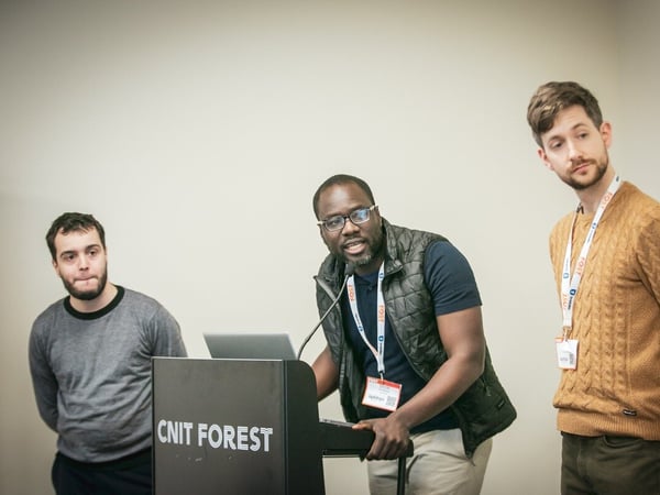 A candid photograph captures three men standing near a black lectern during an indoor presentation or conference, with the central figure speaking into a microphone.