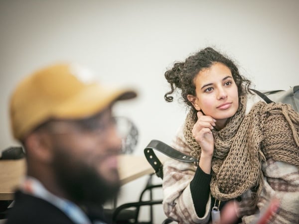 A candid, close-up photograph captures a young woman wrapped in a chunky knit scarf and shawl, looking attentively off-camera, while a blurry man in a yellow cap occupies the foreground.