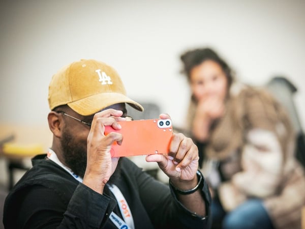 A close-up photograph shows a man wearing a gold baseball cap and glasses, holding an orange smartphone horizontally to take a picture or video in an indoor setting. A second, heavily blurred person is visible in the background.