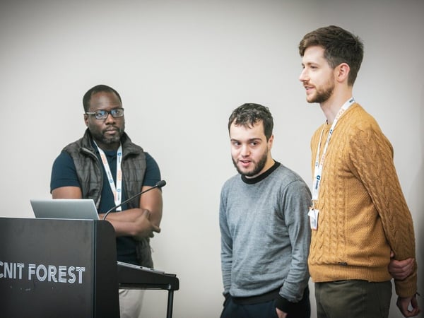 A photograph showing three men standing near a dark podium with a laptop and microphone in an indoor conference or event setting against a plain light wall.
