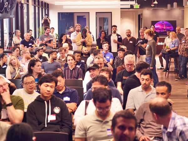 A photograph capturing a large crowd attending an indoor event or presentation, with many people seated and focused toward the front of the room in a modern office setting.