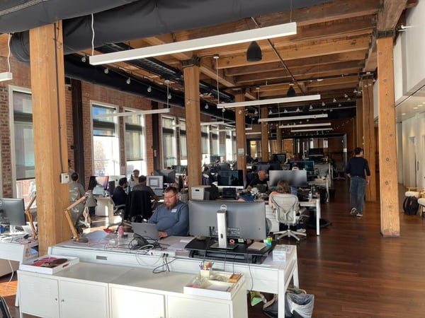 A photograph of a modern, open-plan office space featuring high ceilings with exposed wooden beams and several employees working at their desks.