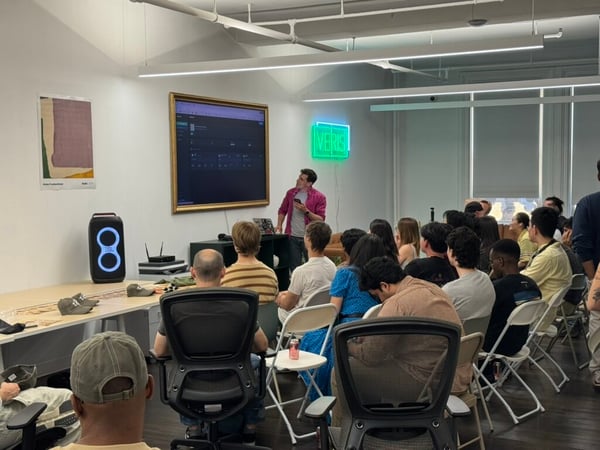 A presenter in a pink shirt stands before a seated audience in an office setting, pointing towards a large screen displaying a software interface. A green neon sign reading 'VERIS' glows on the wall behind him.
