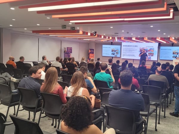 A group of people are seated in a modern conference room, watching a speaker give a presentation in front of several large screens.