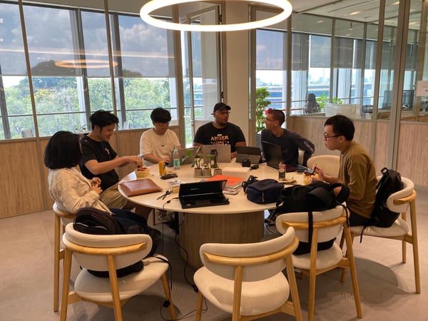 A group of colleagues are gathered around a large circular table in a modern office space, engaged in a mix of work and casual conversation while some eat.