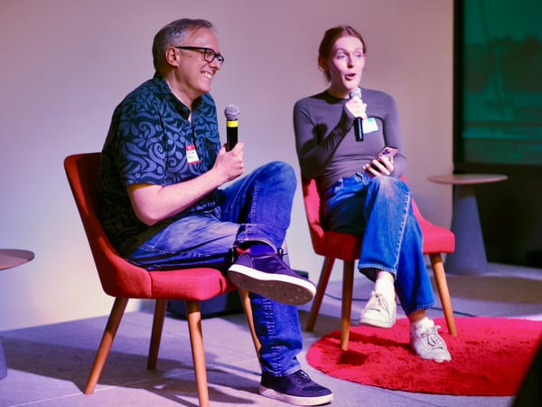 A candid photograph of two people, a man and a woman, sitting in red chairs on a stage, participating in a panel discussion or interview while holding microphones. The scene is lit with dramatic purple and pink ambient stage lighting, with a large dark screen visible in the background.