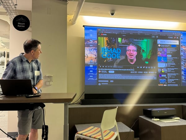 A man stands next to a laptop at a standing desk, looking toward a large projection screen displaying a YouTube video during an indoor presentation or workshop.