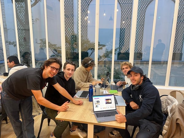 A photograph showing a group of five young men sitting around a wooden table, working on laptops in a bright, indoor space characterized by large arched glass windows and white framing.