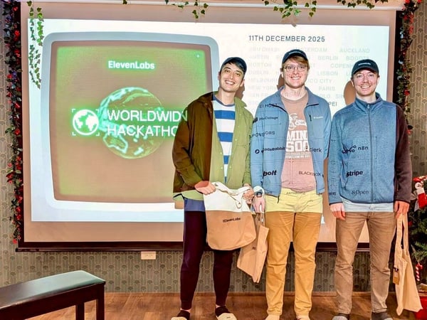 A photo of three young men standing indoors in front of a projection screen that displays the branding for the "ElevenLabs Worldwide Hackathon," along with a date, city list, and sponsor logos. The men are smiling and holding tote bags, likely at the event venue.