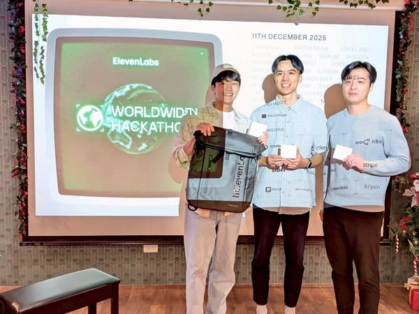 A photograph showing three young men standing indoors in front of a projected screen advertising the ElevenLabs Worldwide Hackathon scheduled for December 11, 2025, while holding prizes including a backpack and small white boxes.