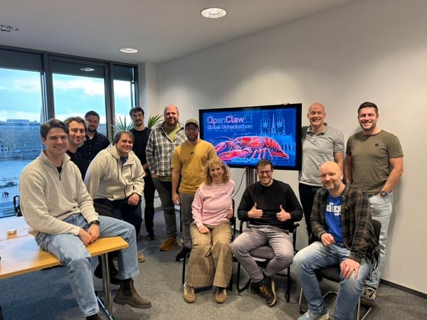 A group of thirteen people are posing for a group photo in an office setting, with a television screen in the background displaying event information.