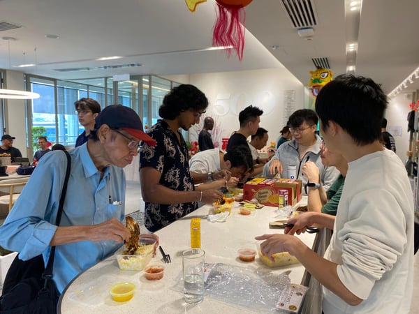 A group of people are gathered around a long white table in a modern indoor space, eating from food containers and socializing. A large '500' logo is visible on the back wall, along with festive decorations like a Chinese lion dance head.