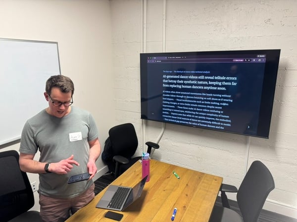 A man stands in an office meeting room, holding a small tablet and appearing to present content displayed on a large wall-mounted monitor that discusses errors in AI-generated dance videos.