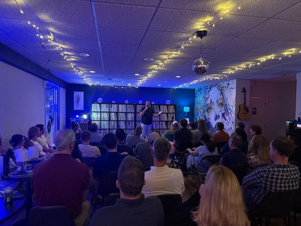 An indoor scene showing a speaker standing at the front of a dimly lit room with blue ambient lighting, addressing an audience seated in rows of chairs.
