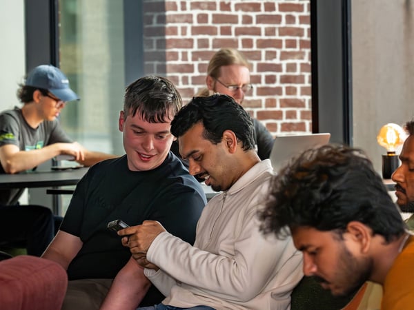 A group of men in a casual indoor setting, with two individuals in the center smiling and looking at a smartphone screen.