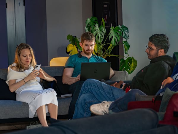 A candid photograph of three people sitting on sofas in a modern lounge or office environment, with one woman looking at her phone, one man using a laptop, and another man looking on.