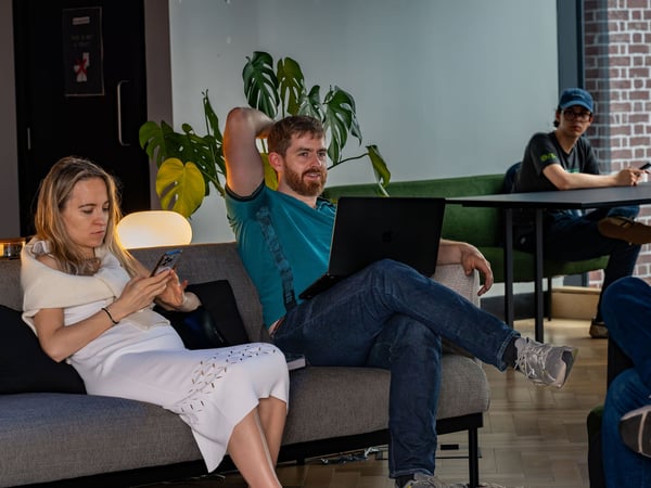 A woman in a white dress looks at her phone while sitting on a sofa next to a man with a laptop in a modern co-working space or lounge.