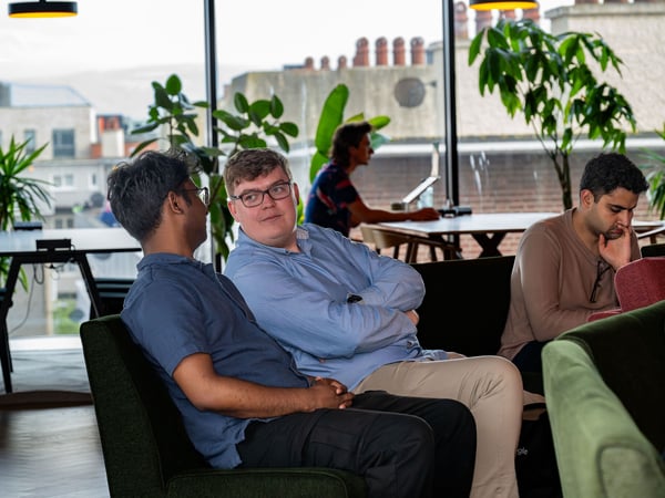 A group of men are seated in a modern, well-lit office lounge with large windows displaying a city view and indoor plants in the background.