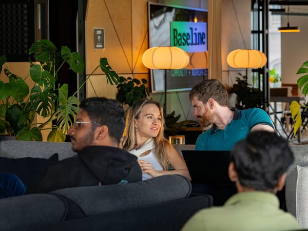 A group of people sitting and talking in a modern, cozy co-working lounge decorated with Monstera plants and warm lighting, with a sign in the background reading 'Baseline COMMUNITY + WORKSPACE'.