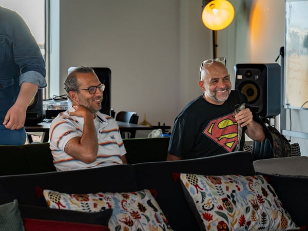 A photograph showing two men sitting on a couch and smiling during an indoor gathering or meeting, with a third man partially visible standing on the left.
