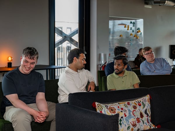 A group of men are seated on couches in a modern office lounge, seemingly listening to a presentation or participating in a meeting.
