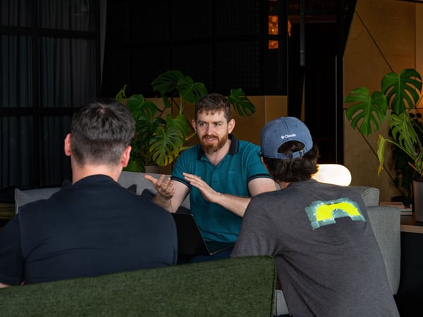 Three men are engaged in a conversation in a modern office lounge setting, with one bearded man gesturing while speaking and the other two listening with their backs to the camera.