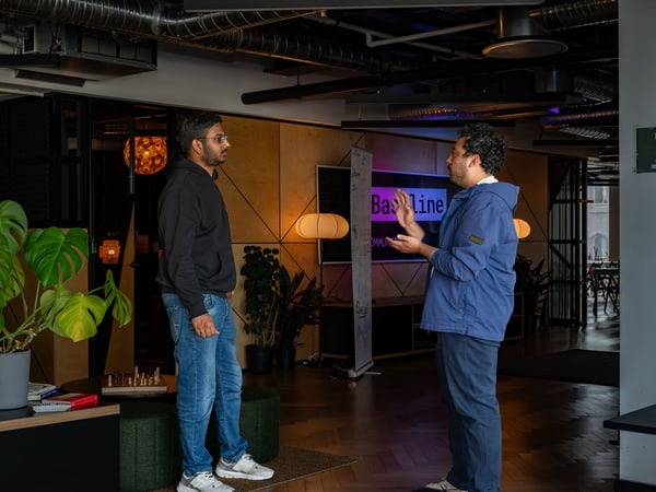 Two men are engaged in a conversation inside a modern office space, with one gesturing as he speaks and the other listening attentively.