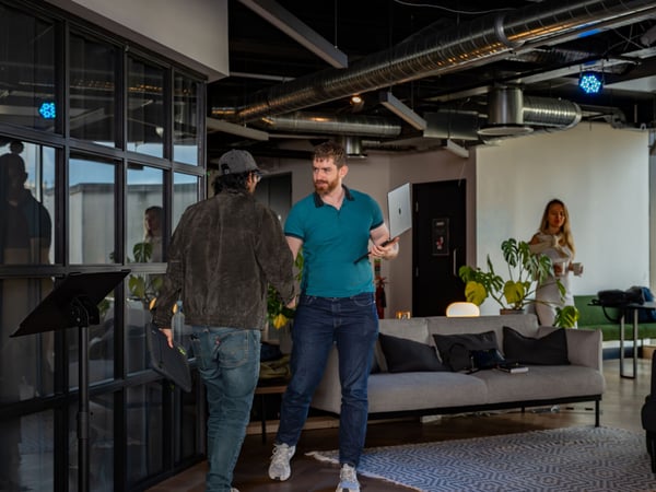 A photograph of a modern office interior where two men are interacting in the foreground, one holding a laptop, while a woman stands in the background near a sofa.