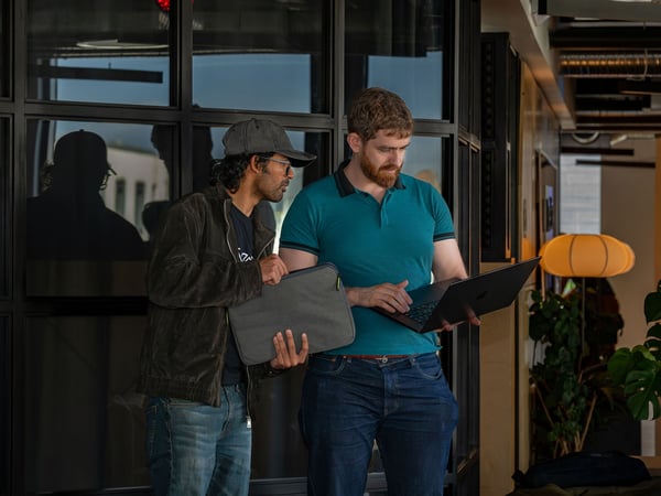 Two men standing in an office environment, looking together at a laptop screen held by one of them.