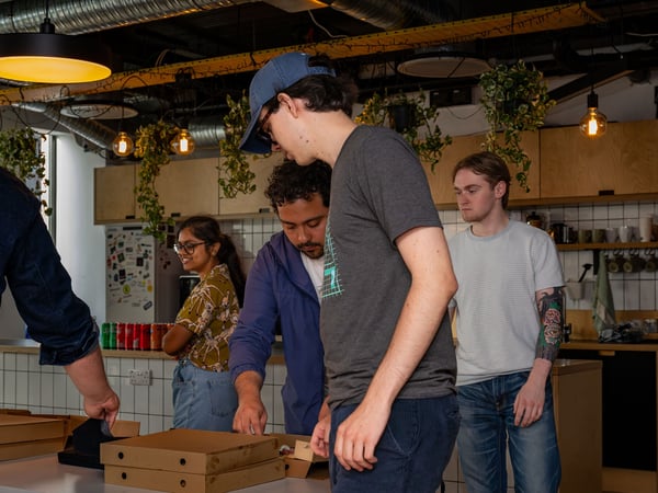 A group of coworkers gathered around a table in an office kitchen, opening pizza boxes during a social gathering.