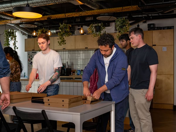 A group of coworkers gathered around a table in an office kitchen, opening several cardboard pizza boxes during a social lunch break.